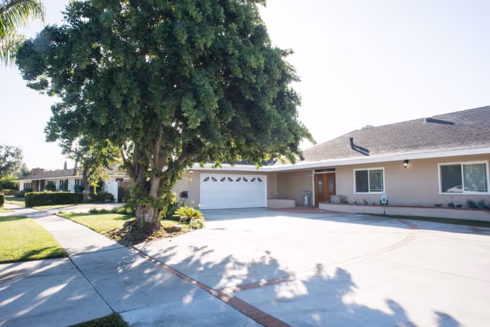 A single-story residential building with a large driveway and a two-car garage. There is a large tree casting shadows on the driveway, and a sidewalk runs along the front lawn. The building has beige exterior walls, white trim, and two front doors with oval glass panels.