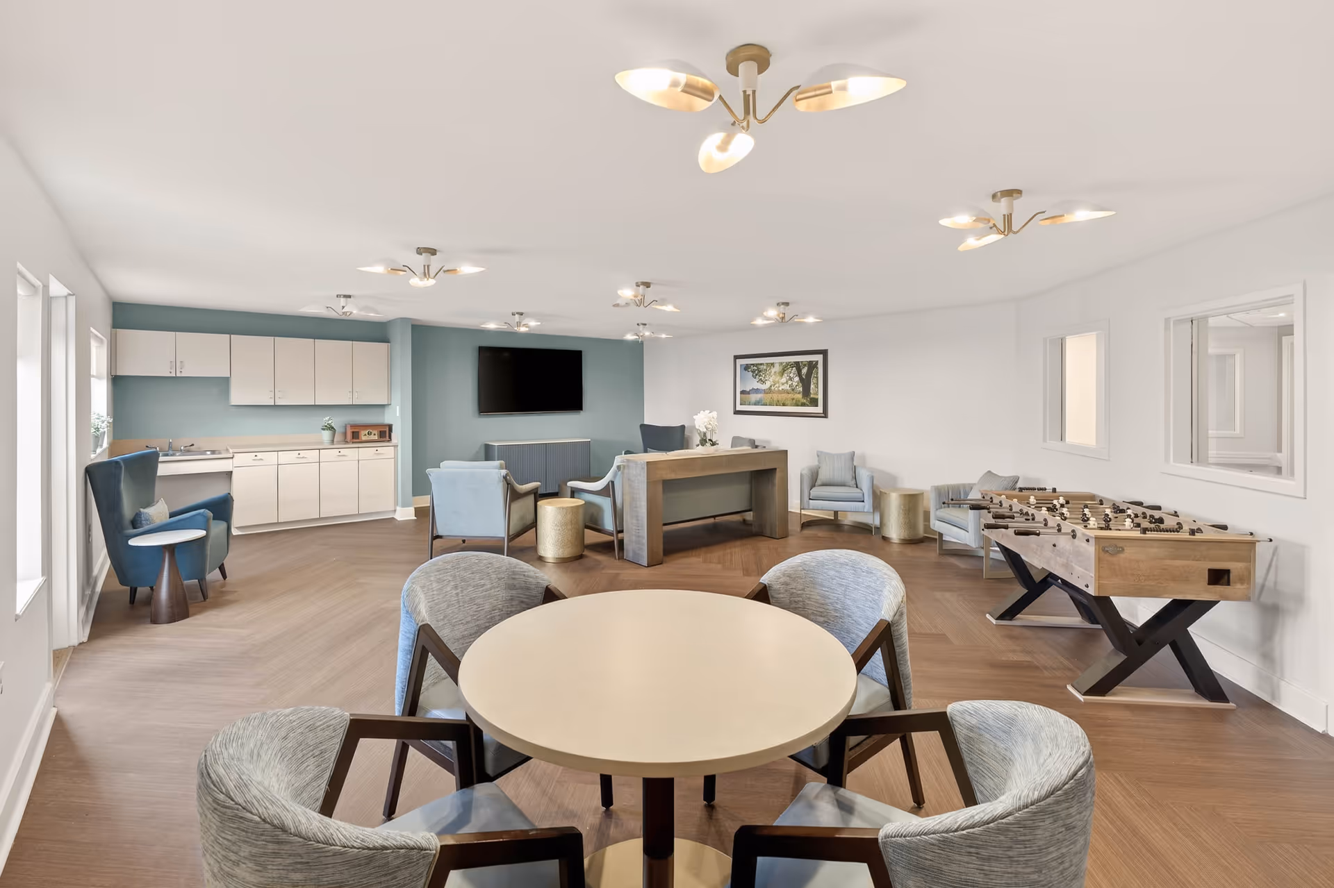 Bright senior living common area with round table, sofas, kitchenette, and foosball table.