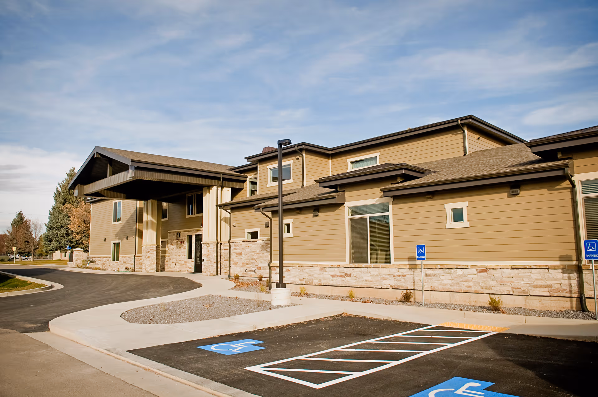 Exterior view of Blacksmith Fork Assisted Living facility showing a modern two-story building with beige siding and stone accents. The entrance has a covered drop-off area, and there are several handicap parking spaces in front. The sky is partly cloudy.