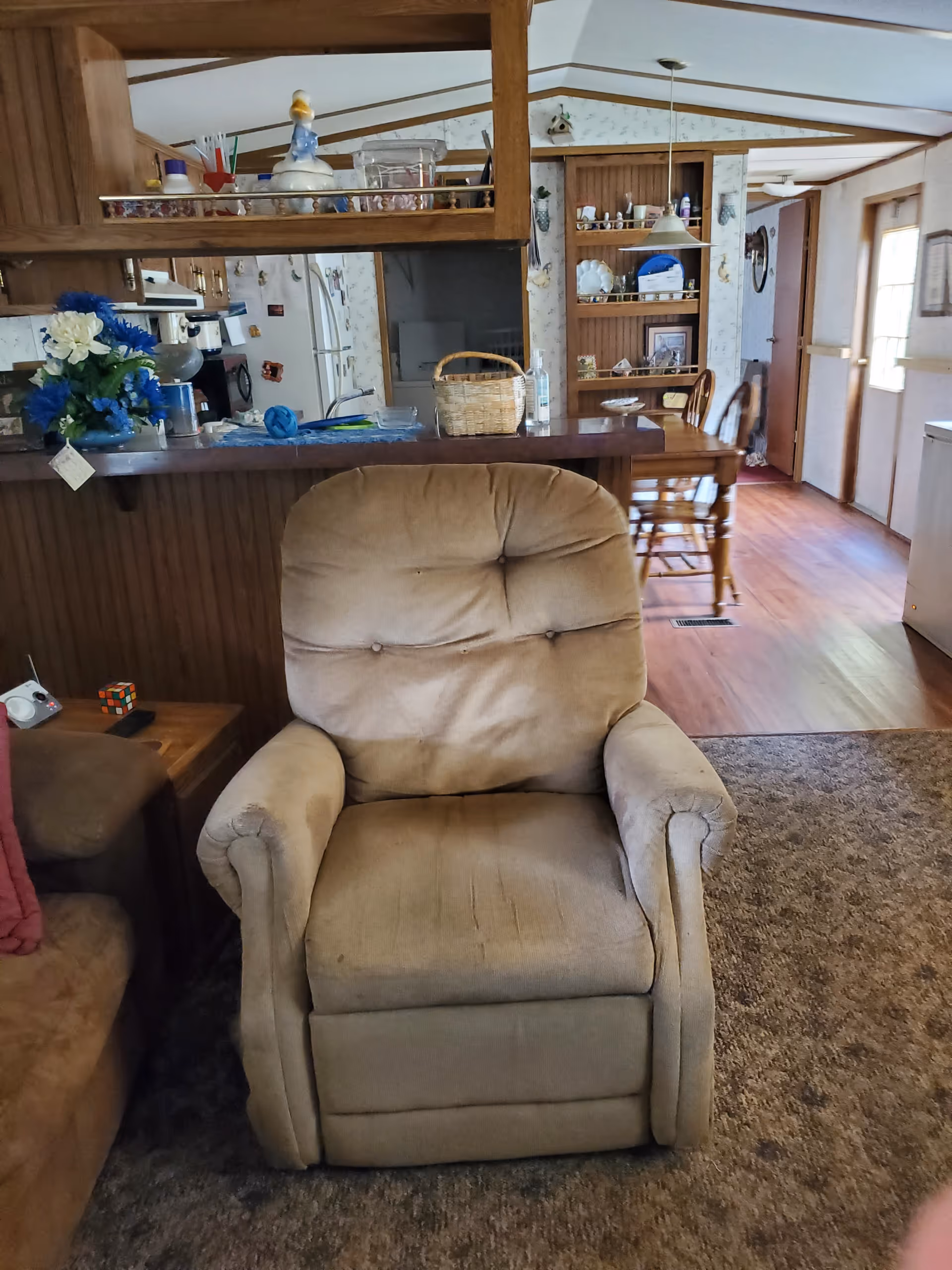 Cozy living room with a beige armchair, carpeted floor, and kitchen visible in background