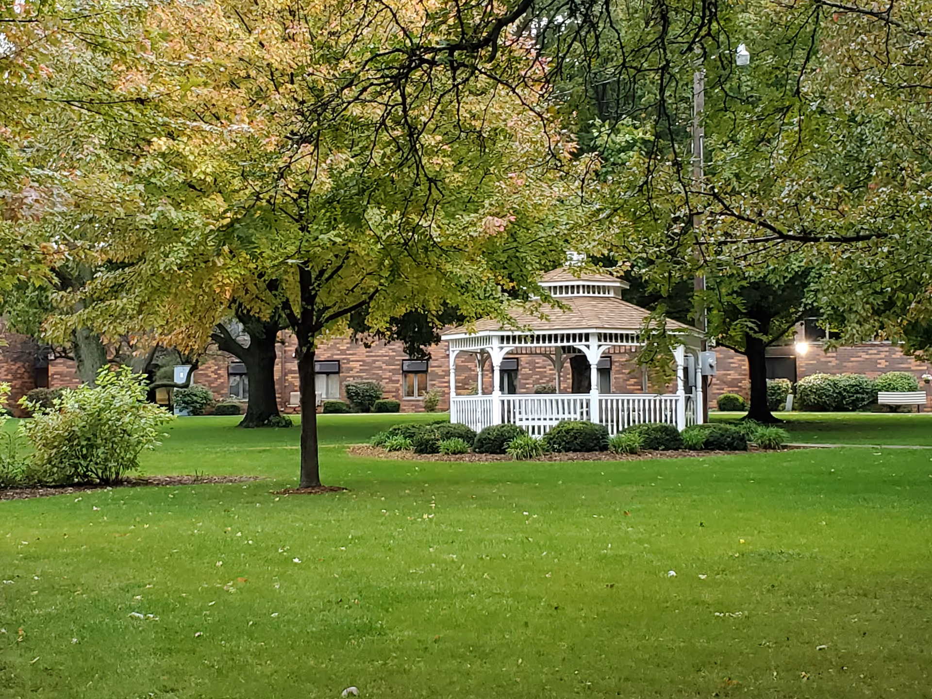 A white gazebo sits on a green lawn surrounded by trees with a brick building in the background.