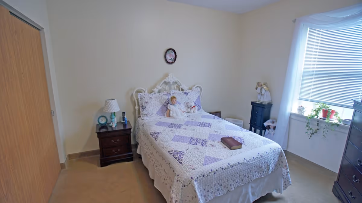 A bedroom with a bed covered in a purple and white patchwork quilt, two dolls placed on the bed, a nightstand with a lamp and clock, a window with blinds and sheer curtains, a small black dresser with angel figurines, and a closet with wooden sliding doors.