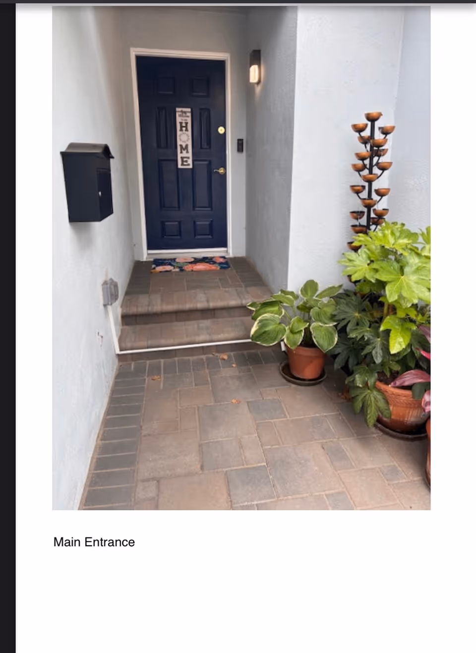 Blue front door labeled Main Entrance; gray walls, potted plants, and a brick pathway.
