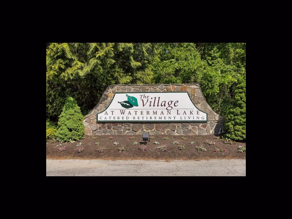 Stone entrance sign reading "The Village at Waterman Lake Catered Retirement Living" with landscaping and trees behind it.