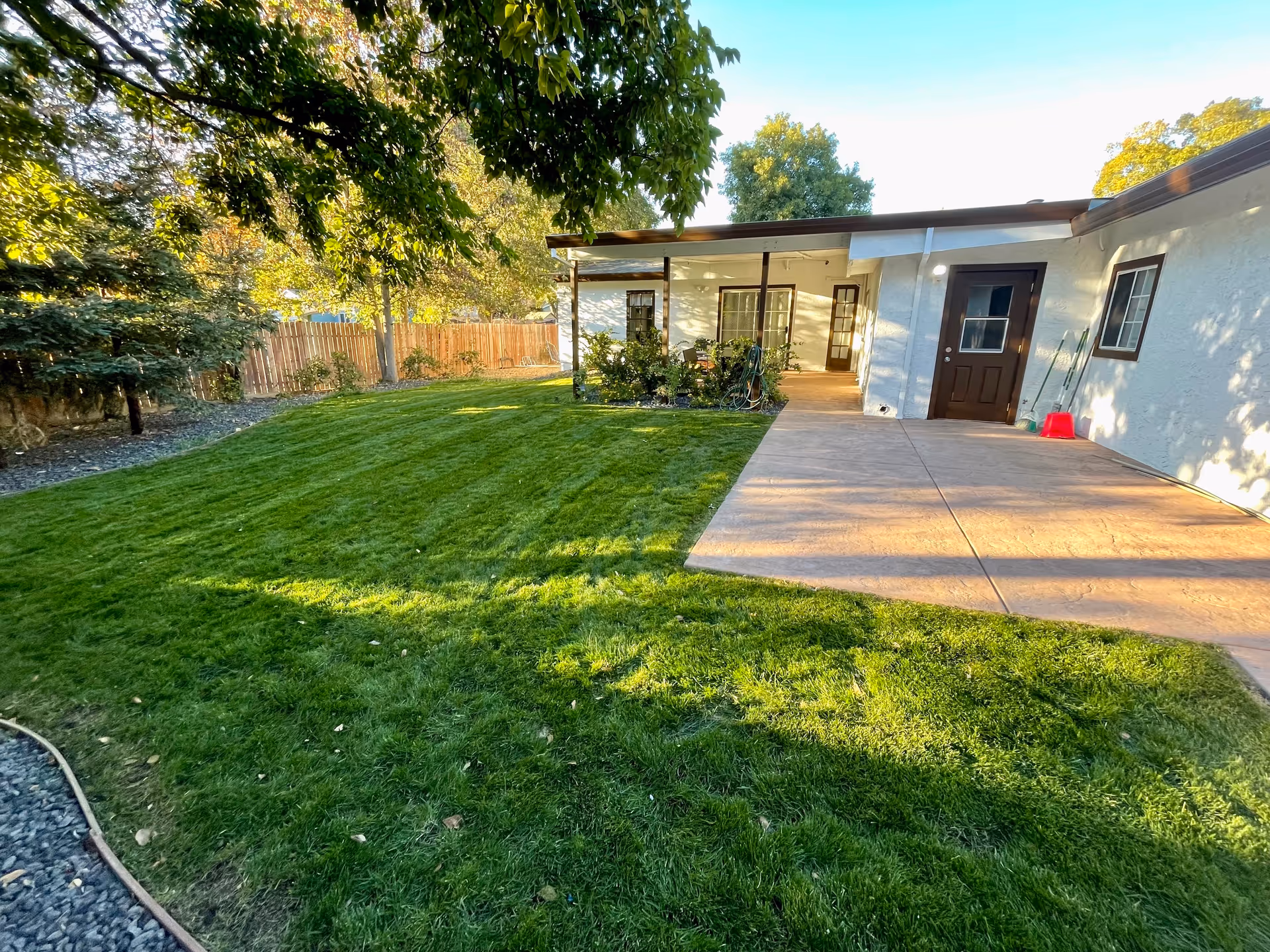 A well-maintained grassy backyard with a covered patio attached to a single-story building, surrounded by trees and a wooden fence.