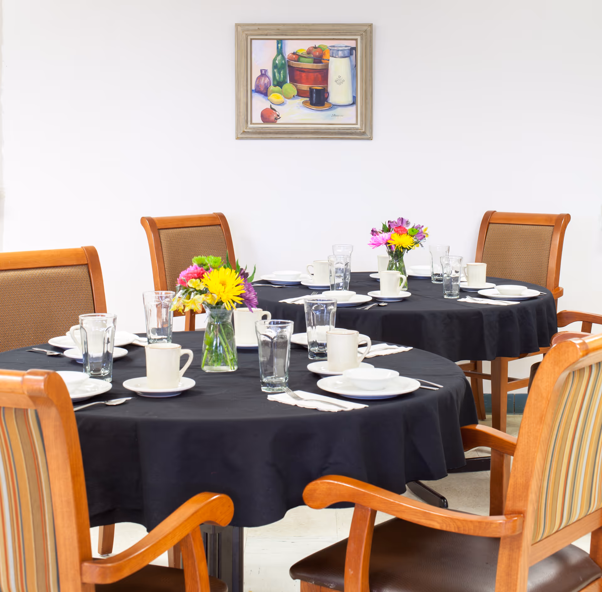 Two round dining tables covered with black tablecloths set with white plates, cups, glasses, and silverware. Each table has a small vase with colorful flowers. Wooden chairs with cushioned seats and backs surround the tables. A framed painting of fruit and bottles hangs on the white wall in the background.