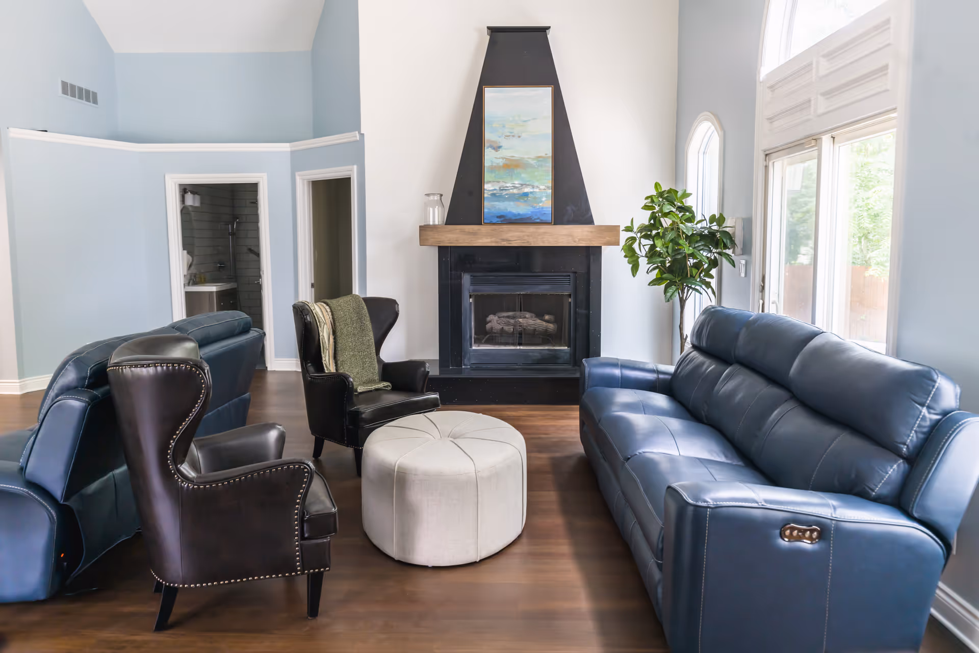 Bright living room with blue leather sofas, chairs, a white ottoman, fireplace, and large window.