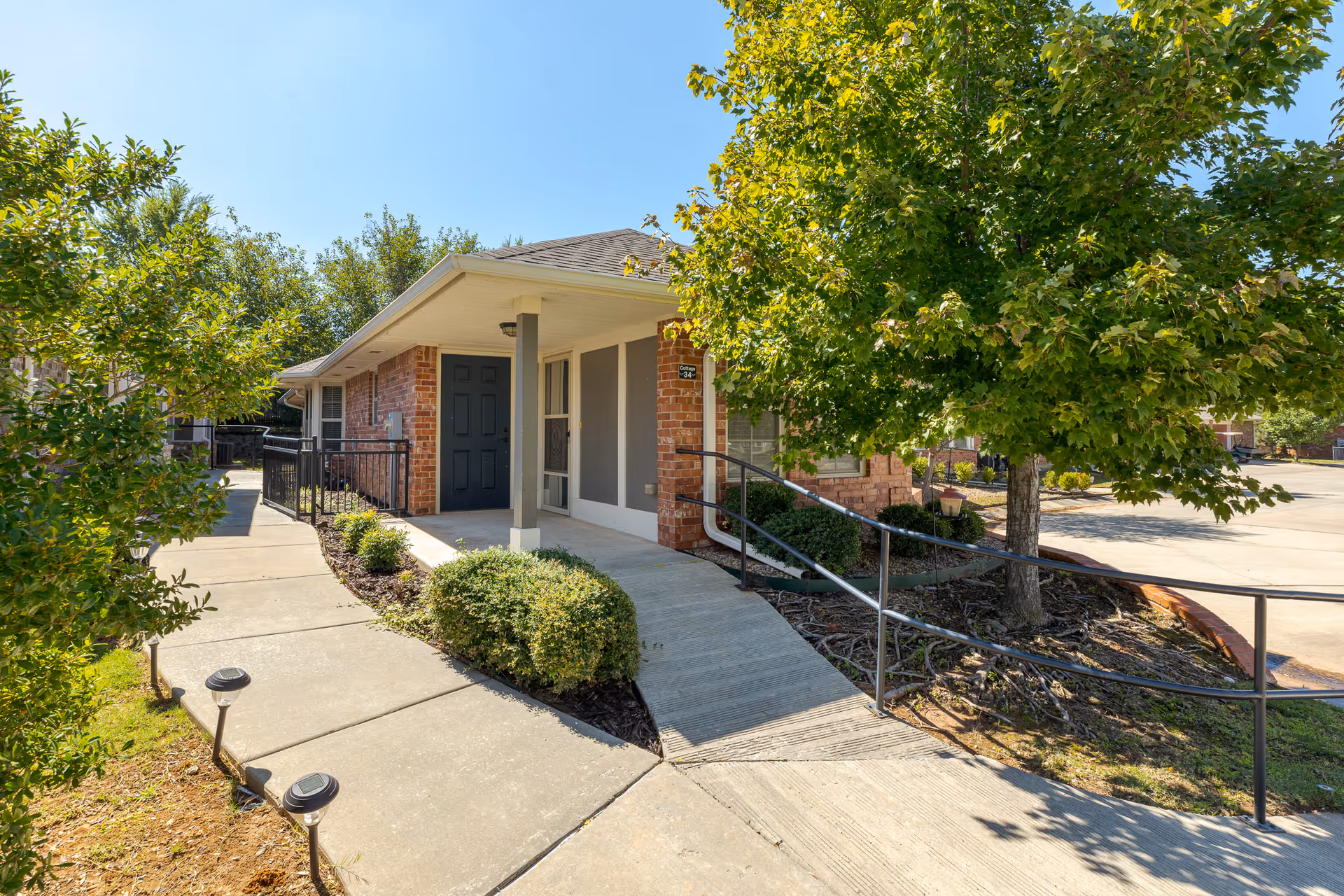 Brick senior living facility entrance with ramp, green shrubs, and trees.