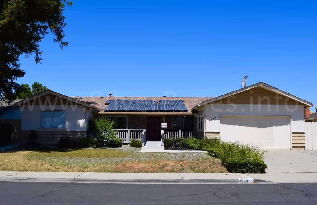 Single-story residential building with a front porch, solar panels on the roof, a garage on the right side, and a lawn with some bushes and plants under a clear blue sky.