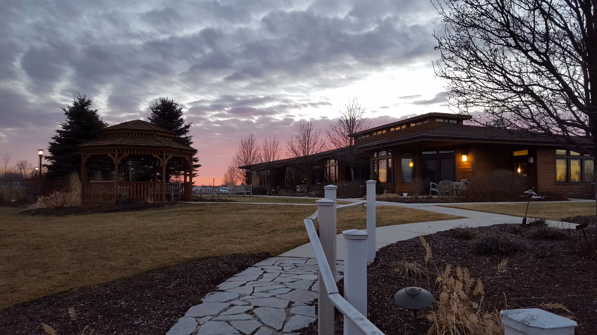 Exterior view of a hospice building with gazebo, path, and sunset.