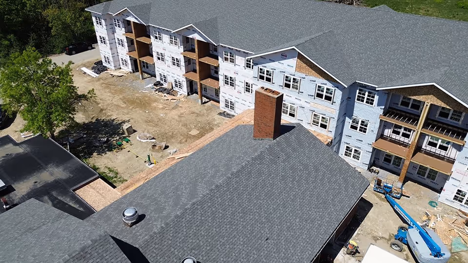 Aerial view of a multi-story building under construction with exposed framing and white Tyvek house wrap on the exterior walls. The building has a gray shingled roof and a large brick chimney. Construction equipment and materials are visible on the ground around the building.