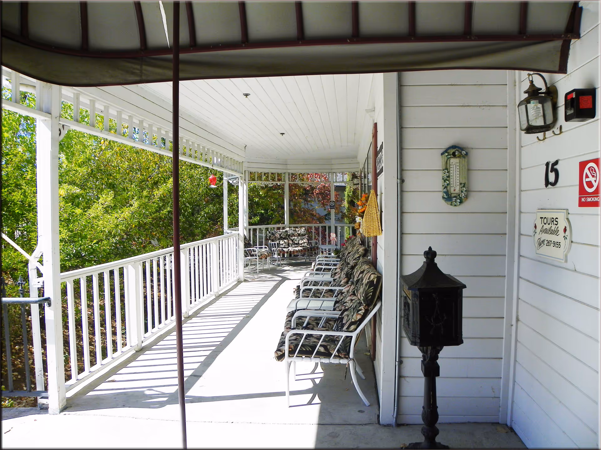 Covered wraparound porch at a senior retirement home with white railings, a row of cushioned chairs, and entrance signage.