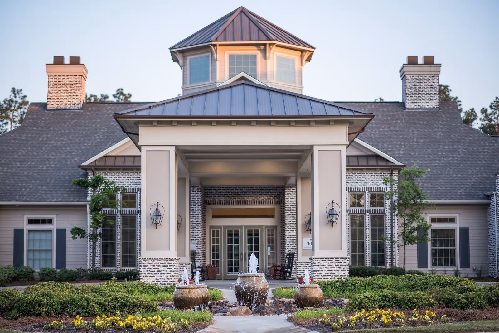 Front exterior view of a senior living facility building with a peaked roof, large windows, two chimneys, a covered entrance supported by columns, and a decorative water fountain in the landscaped garden in front.