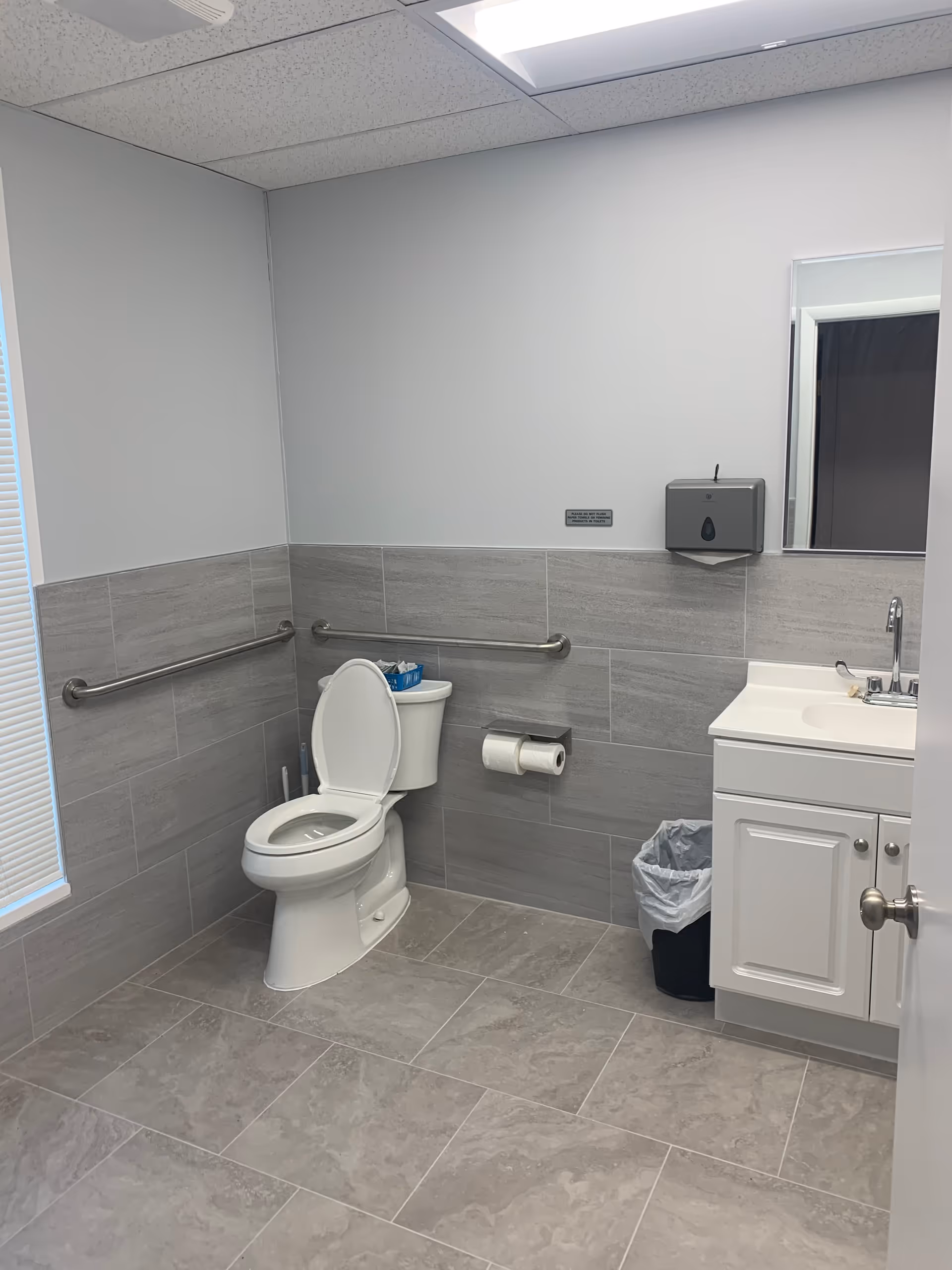 Bright bathroom with toilet, sink, mirror, wall-mounted grab bars, and gray tiled walls.
