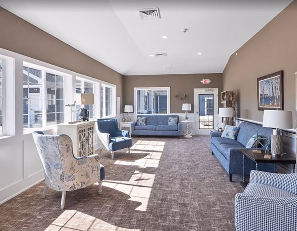 Sunlit communal living room with blue sofas and armchairs arranged along large windows.
