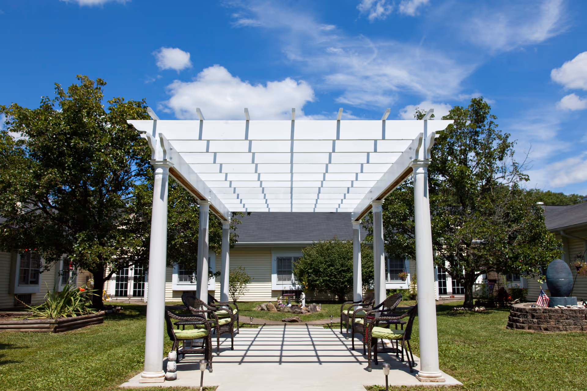 Outdoor seating area with a white pergola casting shadows on the concrete floor, surrounded by green grass, trees, and a building in the background under a blue sky with scattered clouds.