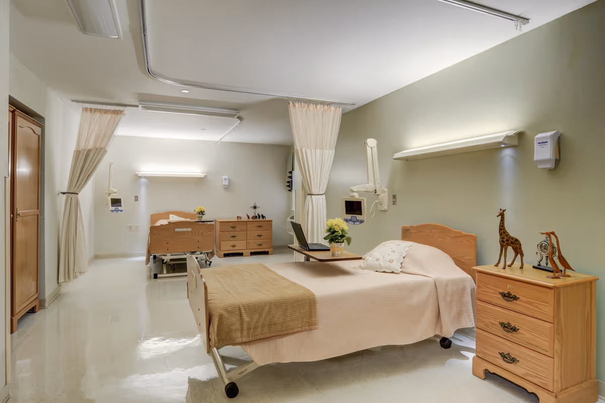 Bright hospital-style patient room with two beds, curtains, and wooden dressers.
