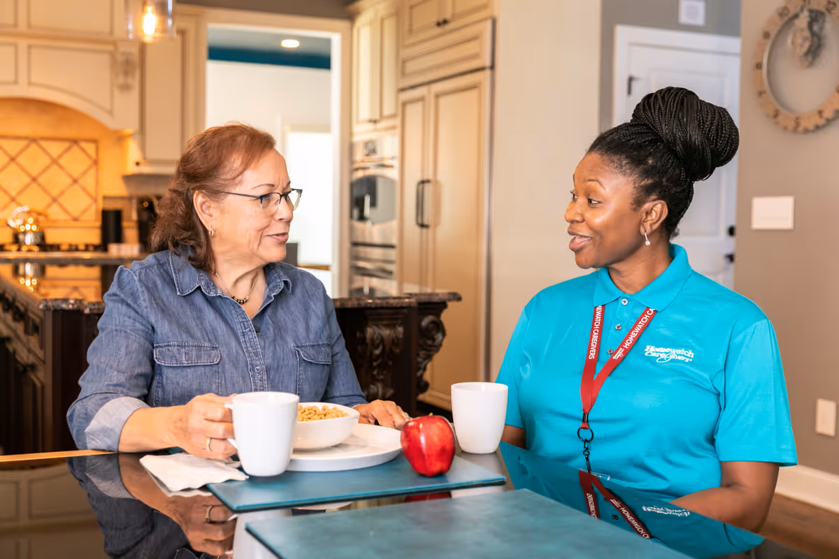 Two women chat at a kitchen island with cups and a bowl of cereal.
