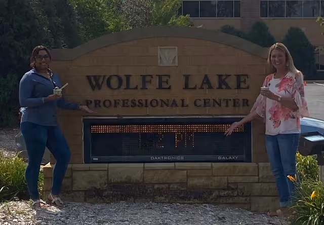 Two women stand beside a Wolfe Lake Professional Center sign outdoors.