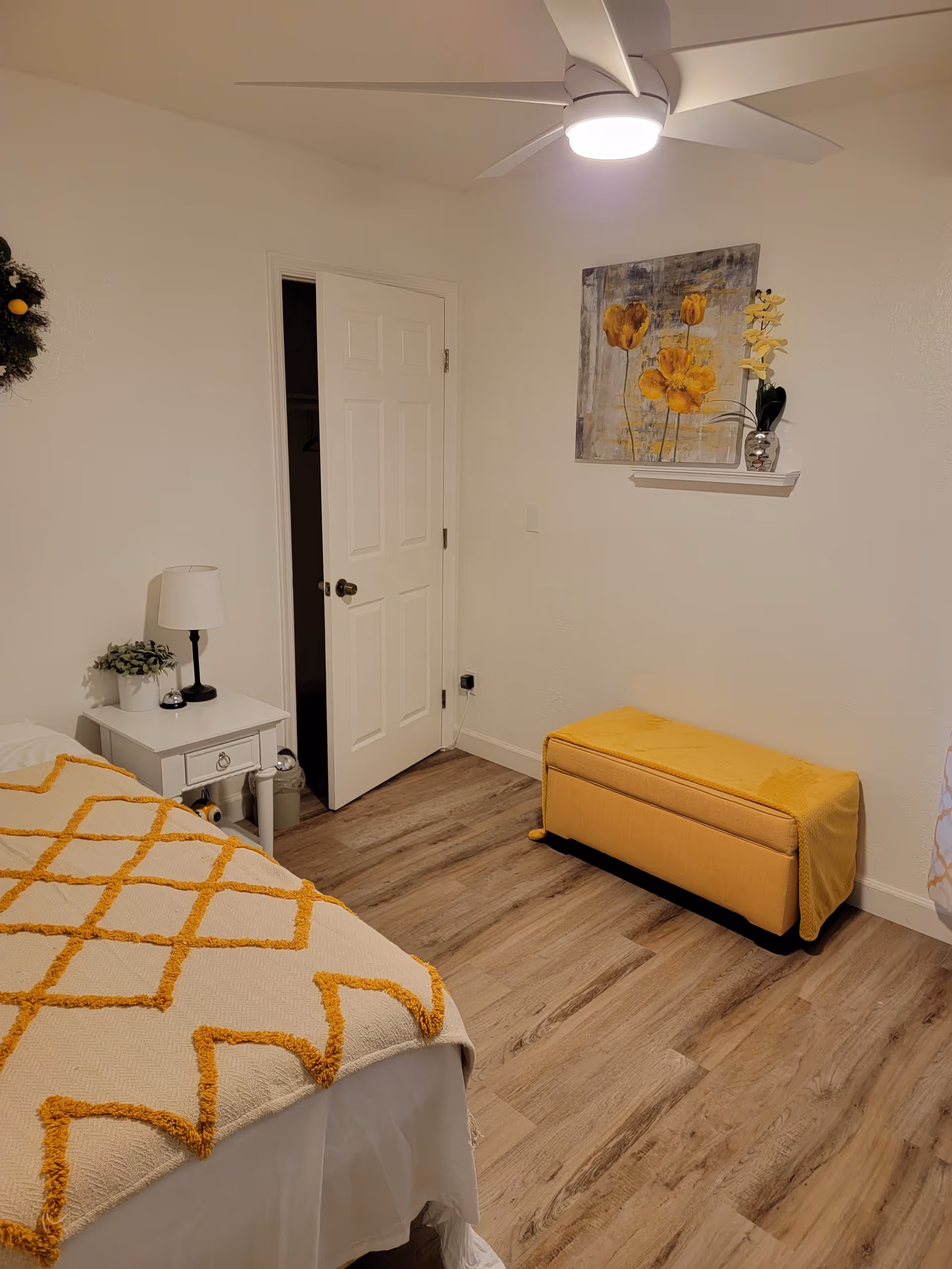Light-filled bedroom with a bed, nightstand, lamp, and yellow ottoman.