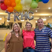 Three adults standing indoors under colorful balloons and silver letter balloons spelling TRHC. The setting appears to be a well-lit common area with plants and mirrors in the background.