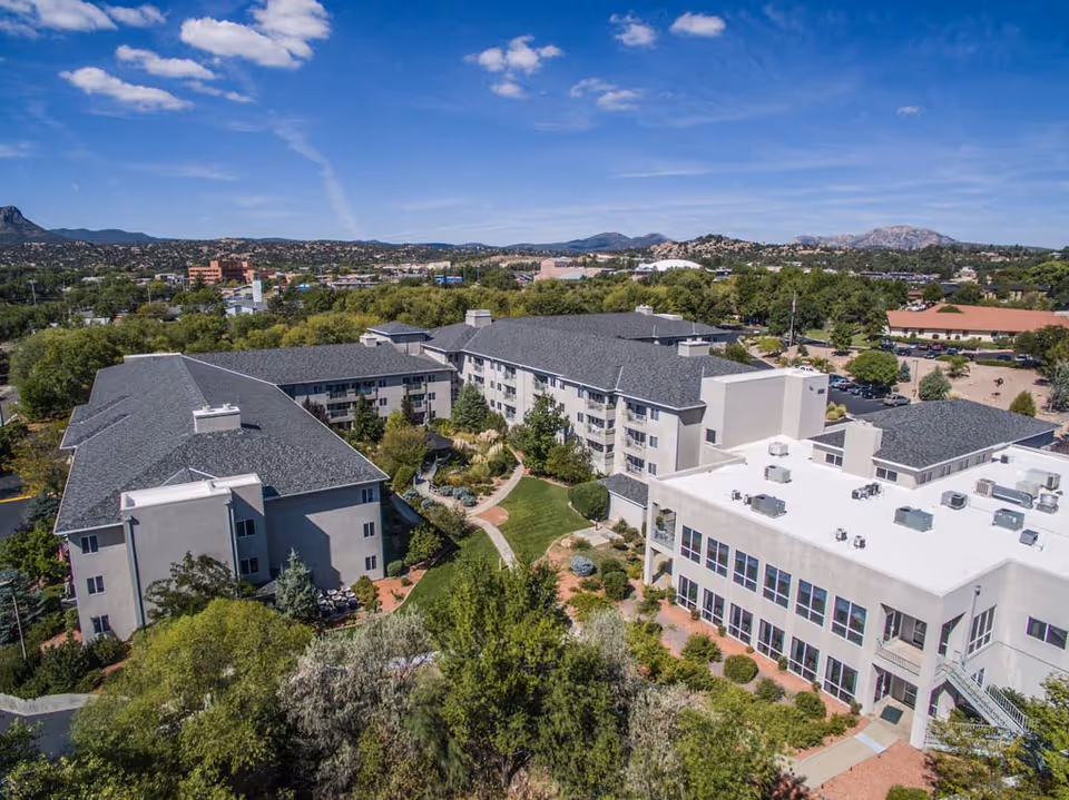 Aerial view of Las Fuentes Resort Village showing multiple connected buildings with gray roofs surrounded by trees and landscaped gardens under a blue sky with scattered clouds. Mountains and a town are visible in the background.