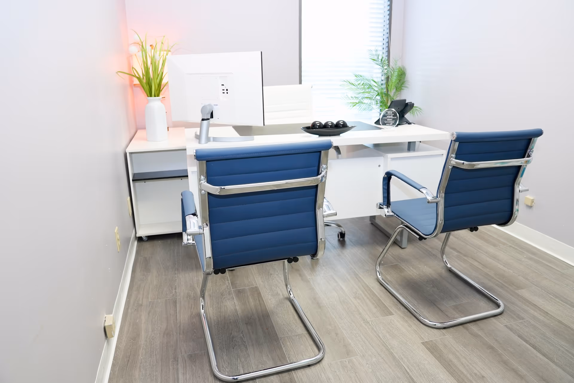 Reception desk with computer, potted plants, and two blue chairs in a bright office.