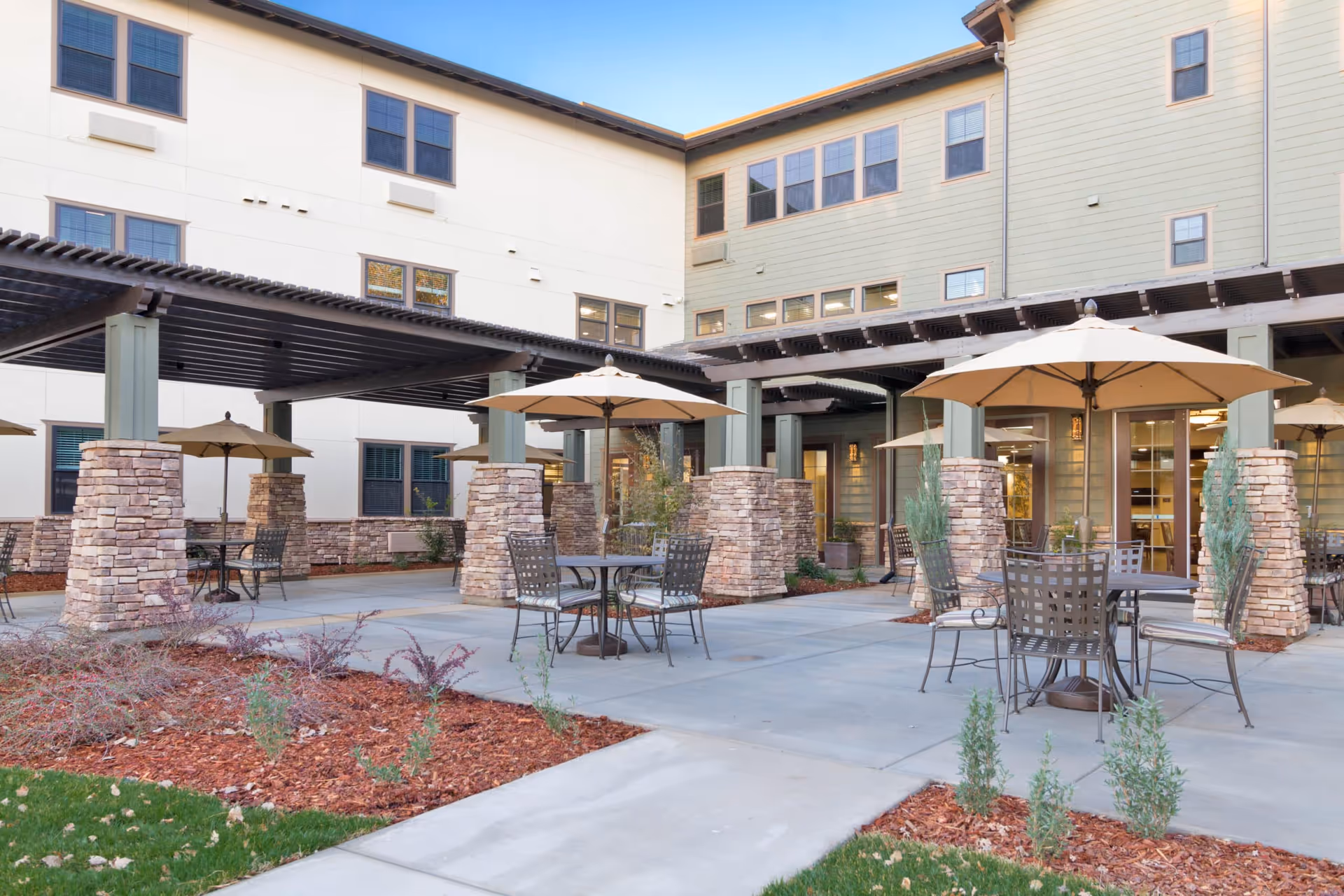 Outdoor patio area at Westmont Of Chico - The Inn featuring multiple round tables with metal chairs and beige umbrellas. The patio is surrounded by a building with multiple windows and stone pillars supporting wooden pergolas. There are landscaped areas with mulch and small plants around the patio.