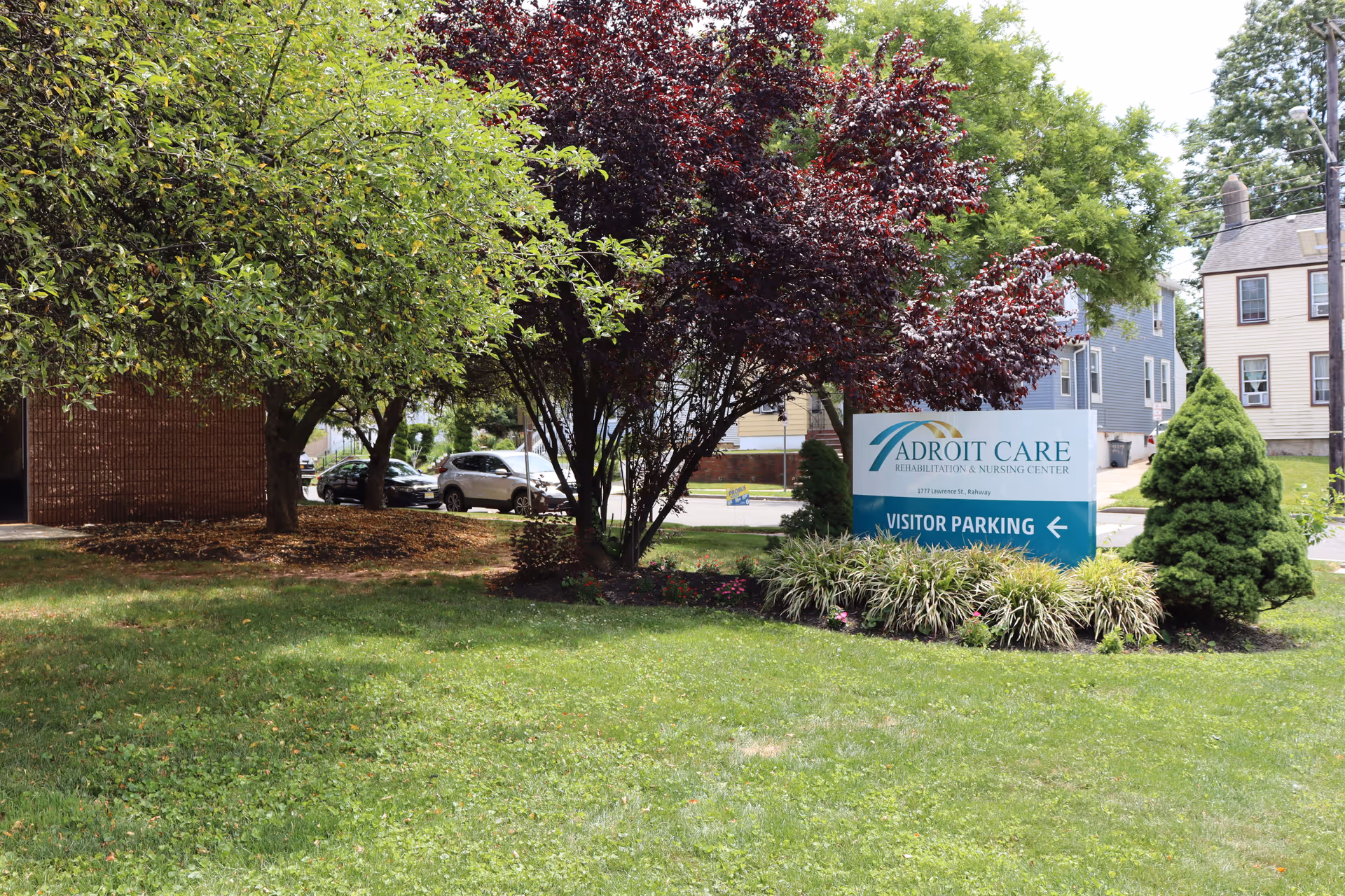 Outdoor grassy area with trees and a blue Adroit Care sign near a residential building.