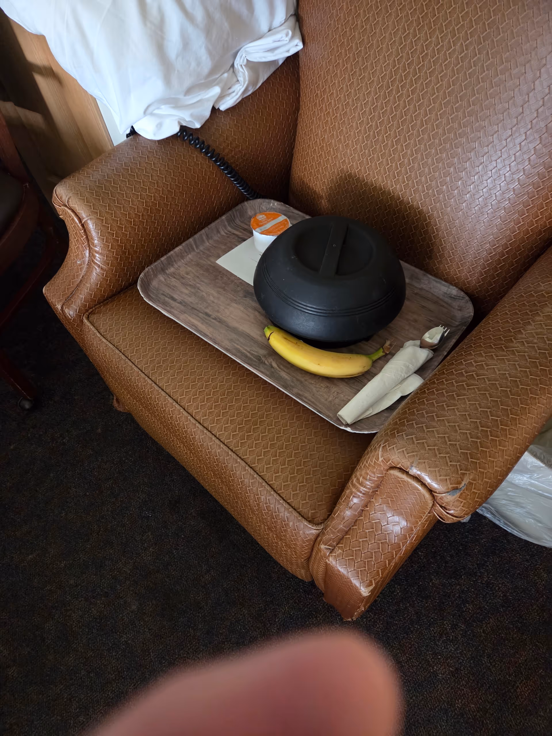 Brown upholstered armchair with a tray holding a banana, napkin, small cup, and black domed lid.