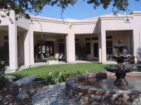 White assisted living building with brick fountain, green lawn, and covered outdoor seating.