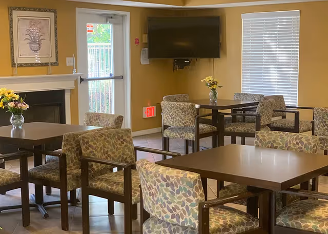 Senior living dining area with tables, patterned chairs, flowers, a wall mounted TV, and windows.