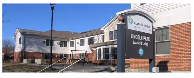 Exterior view of Essentia Health Lincoln Park Assisted Living facility showing a two-story building with white siding and red brick accents. A sign in front displays the facility name and logo. There is a sidewalk and a streetlamp visible in the foreground.