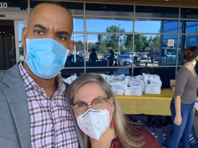 Two masked individuals pose near a table of bags outside Ultimate Home Health Care.