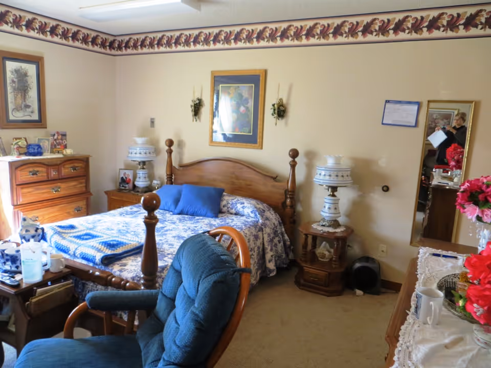 A cozy furnished bedroom with a wooden bed covered in blue patterned bedding, bedside tables with lamps, a dresser, an upholstered chair, and wall art.