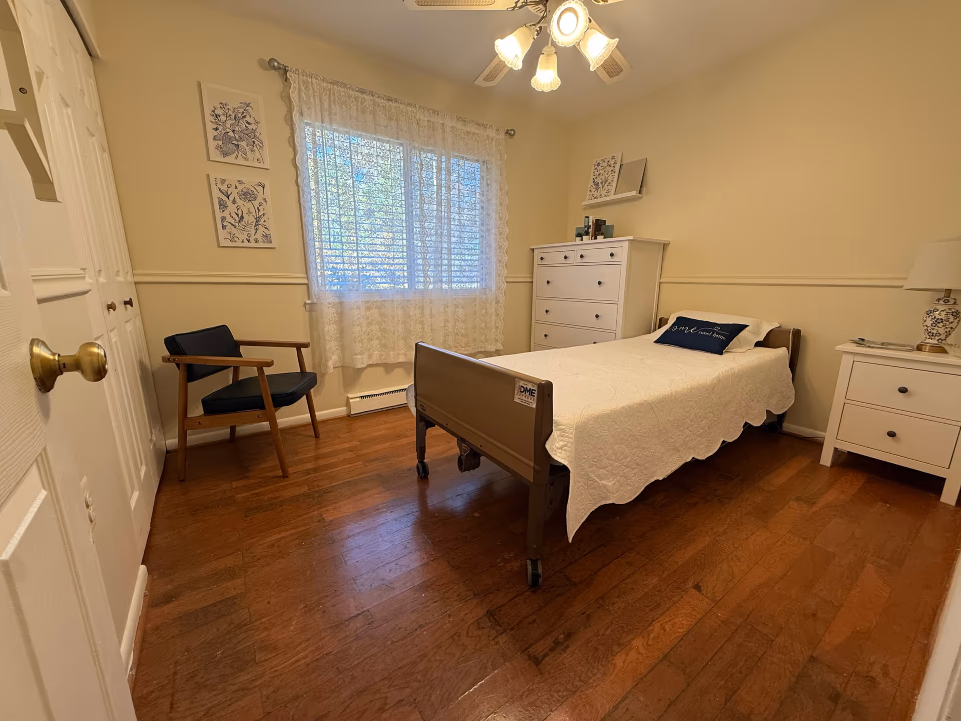 A cozy bedroom with bed, dresser, chair, and lace-curtained window.