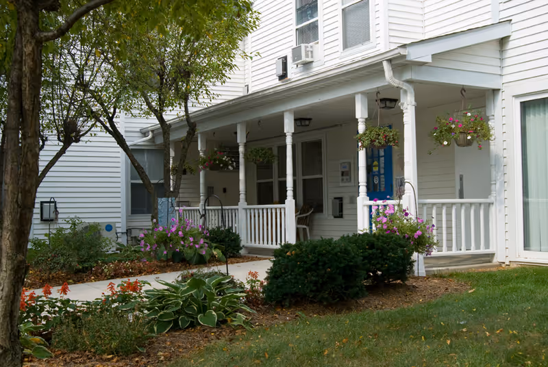 A white building with a covered porch featuring white railings and columns. The porch has hanging flower baskets with pink flowers and a blue door. There are bushes, flowering plants, and a tree in the landscaped garden area in front of the porch.