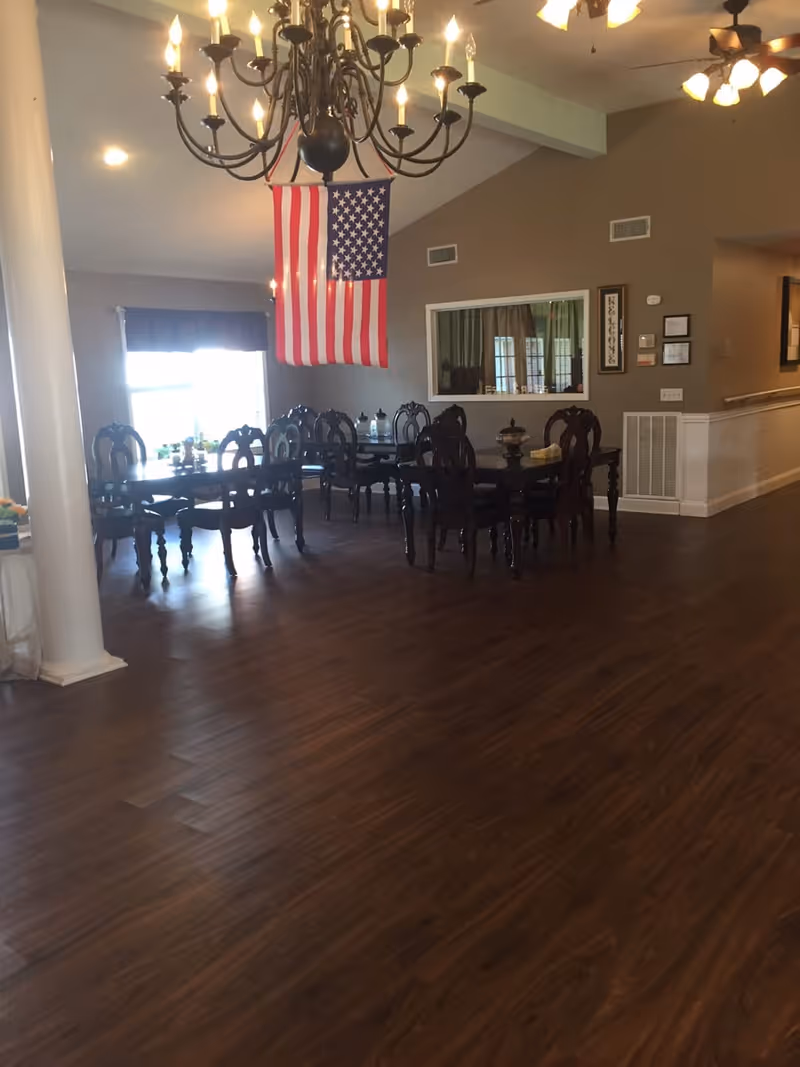 Interior view of a dining area with dark wooden tables and chairs arranged neatly on a polished wooden floor. An American flag hangs from the ceiling near a large chandelier with multiple lit bulbs. The room has beige walls, a window with a curtain, and a ceiling fan with lights.