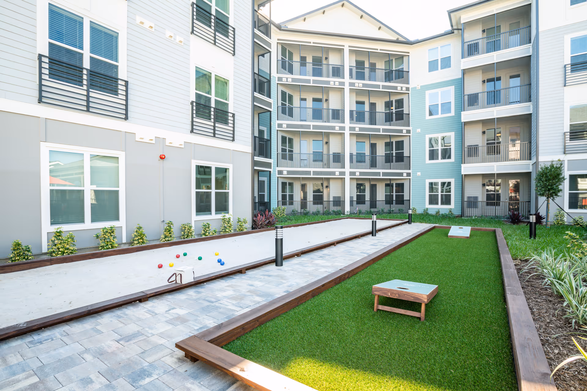 Outdoor courtyard area of a senior living facility featuring bocce ball and cornhole game setups, surrounded by a multi-story building with balconies and windows.
