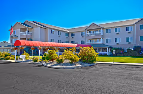 Front exterior of a multi-story senior living facility with a red covered entrance, landscaped shrubs, and an American flag.