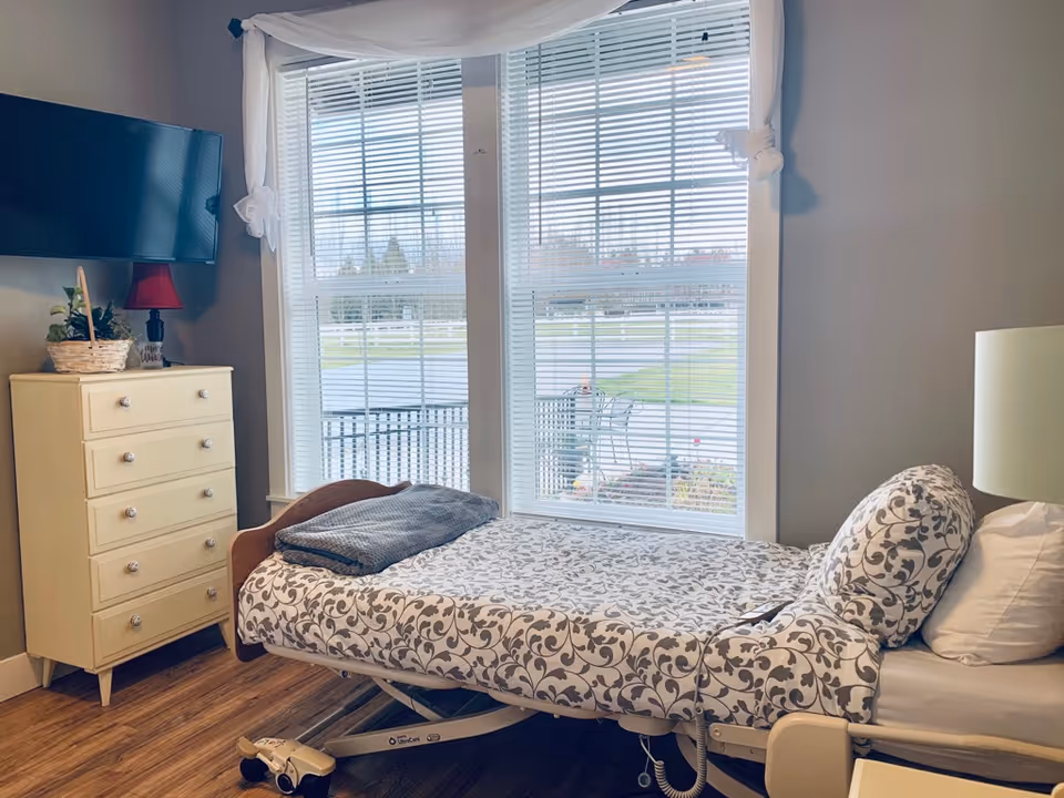 Senior living bedroom featuring an adjustable bed, dresser, TV, and large window with blinds.