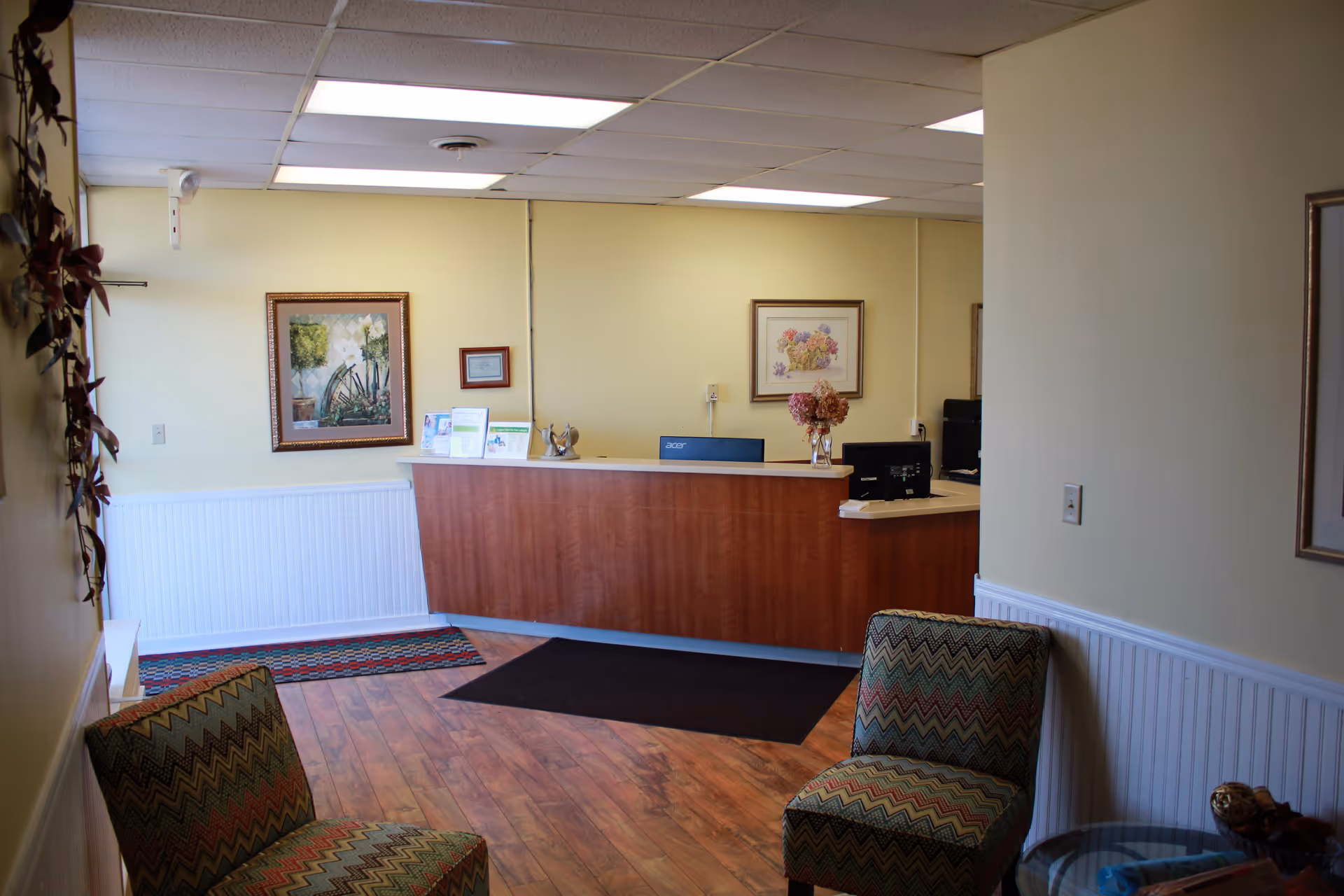 Reception desk with computer, floral arrangement, framed art, and two patterned chairs in a lobby.