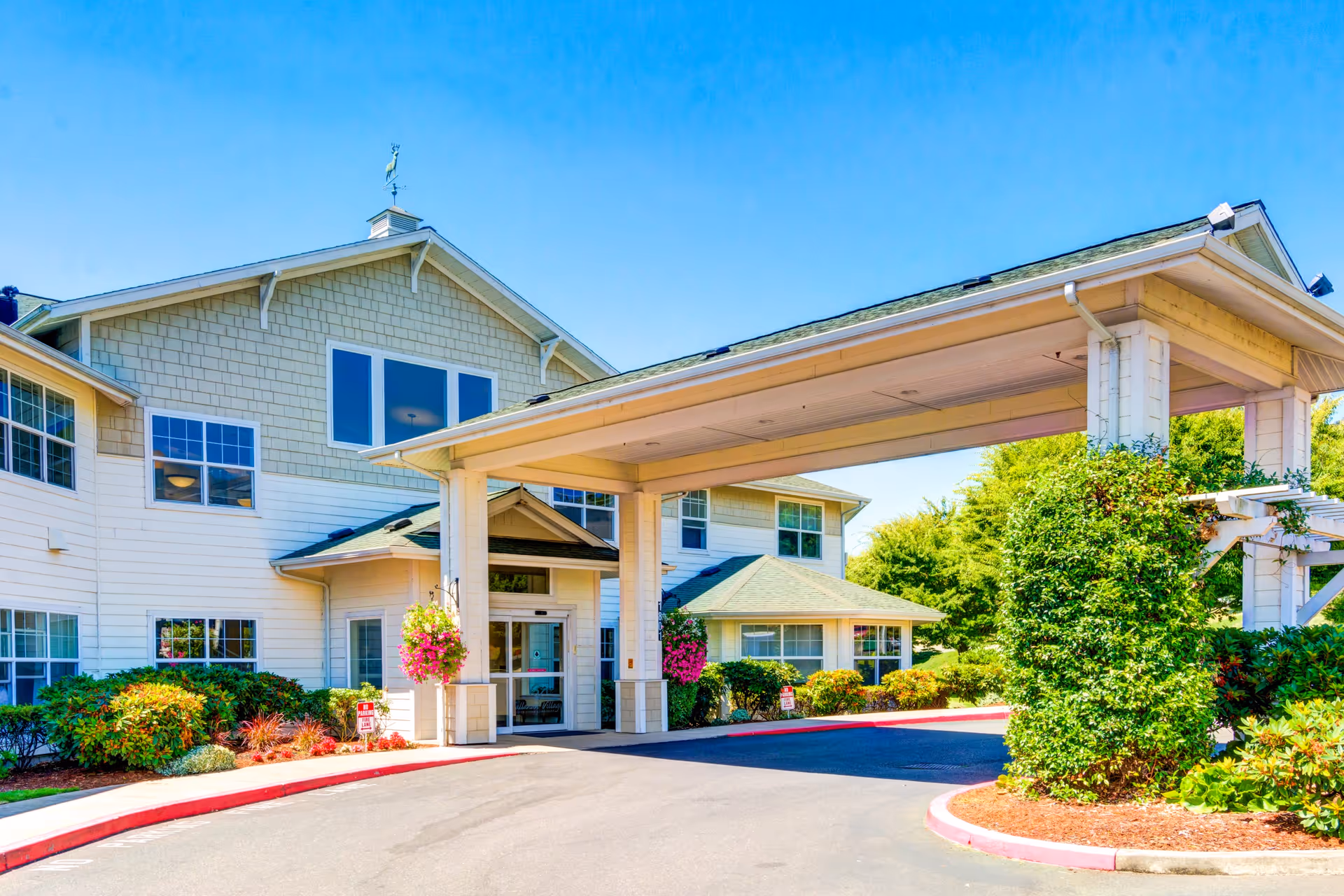 Front entrance of a retirement and assisted living building with a covered drop-off canopy, hanging flower baskets, and landscaped grounds.