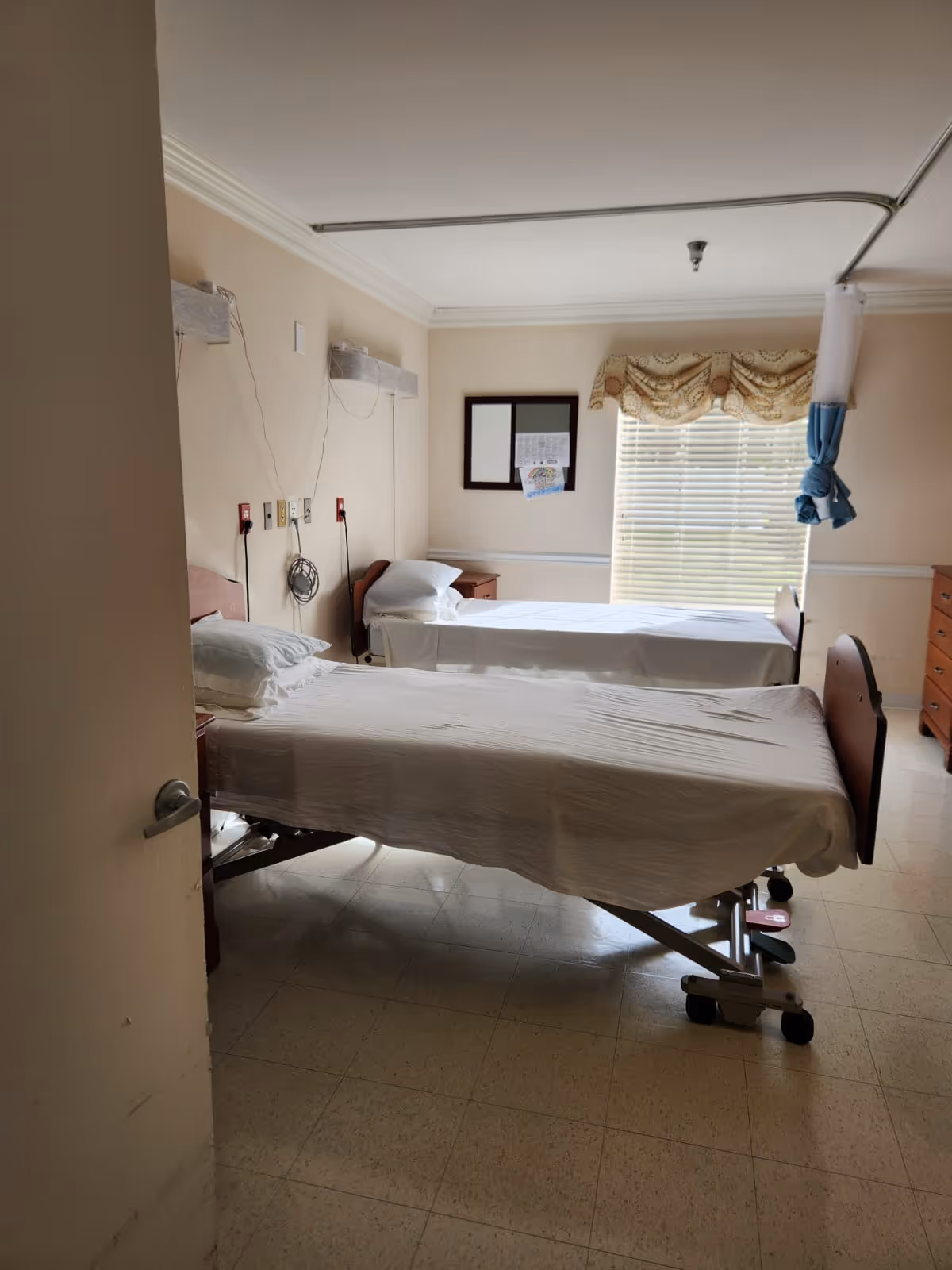 Two hospital beds in a patient room with window blinds and medical equipment.
