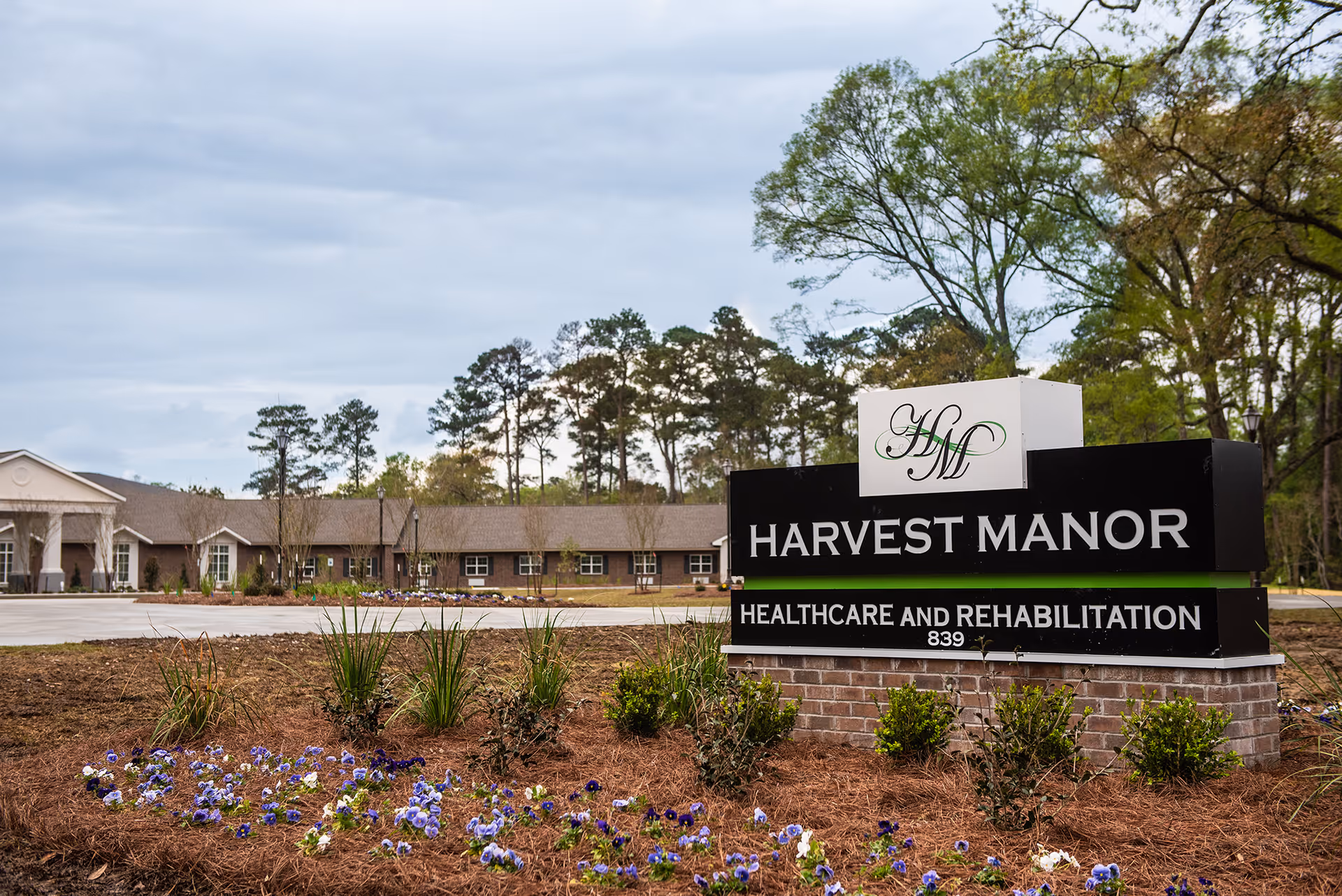 Exterior view of Harvest Manor Healthcare and Rehabilitation building with a large sign in the foreground displaying the facility's name. The building is surrounded by trees and landscaped with flowers and shrubs.