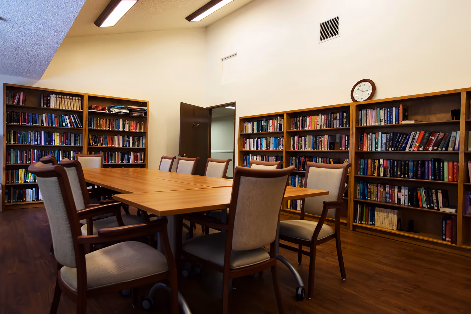 A quiet room with wooden bookshelves filled with books along the walls, a large wooden table in the center surrounded by cushioned chairs with wooden frames, and a clock on the wall above the bookshelves.