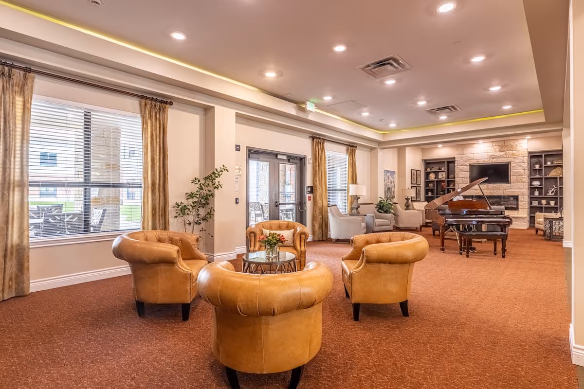 A spacious and well-lit living room area in an assisted living facility featuring four tan leather armchairs arranged around a small glass coffee table with a flower arrangement. The room has large windows with gold curtains, a grand piano near a stone fireplace, built-in shelves with decorative items, and additional seating with light-colored armchairs and lamps.