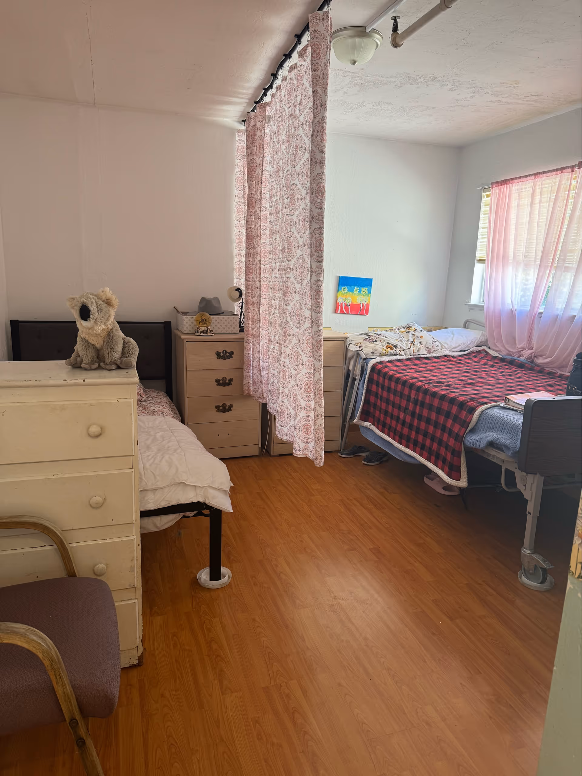 Shared senior bedroom with pink curtain divider, two beds, dresser, and teddy bear.