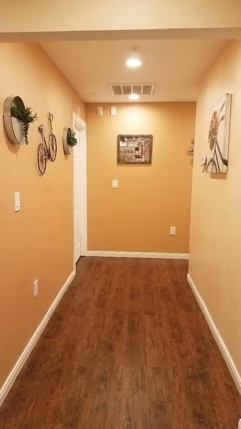 A narrow hallway with wood flooring, beige walls, recessed ceiling lights and decorative wall art leading to a closed door.
