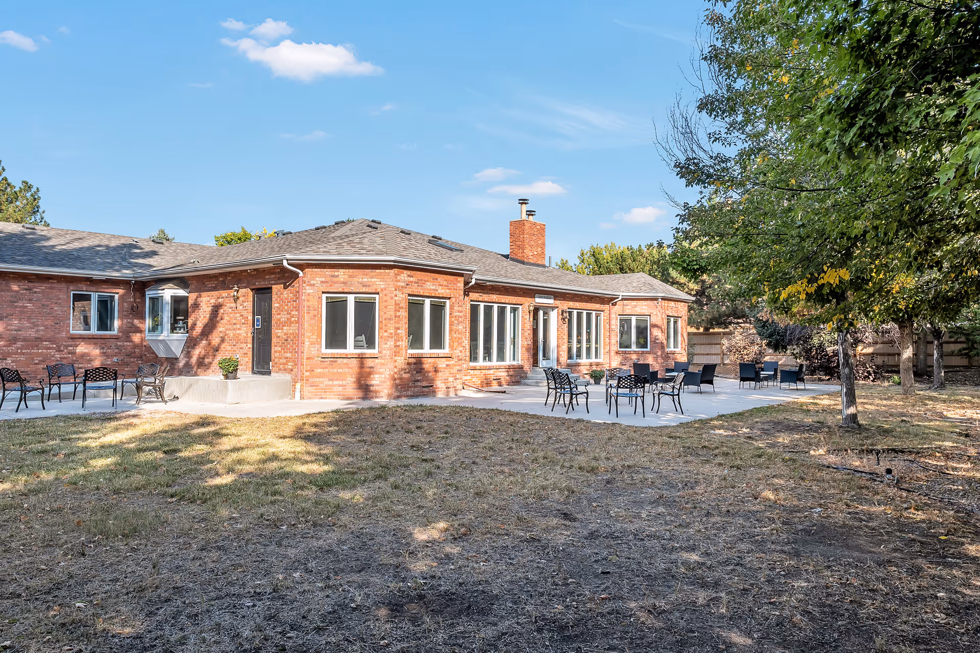 Outdoor view of a single-story brick building with multiple windows and a chimney, surrounded by a patio area with several black metal tables and chairs. There are trees and a grassy yard in the foreground under a blue sky with a few clouds.